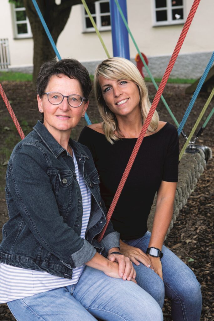 Zwei Frauen auf einem Spielplatz, sitzen auf einer Seilwagen, umgeben von bunten Seilen.