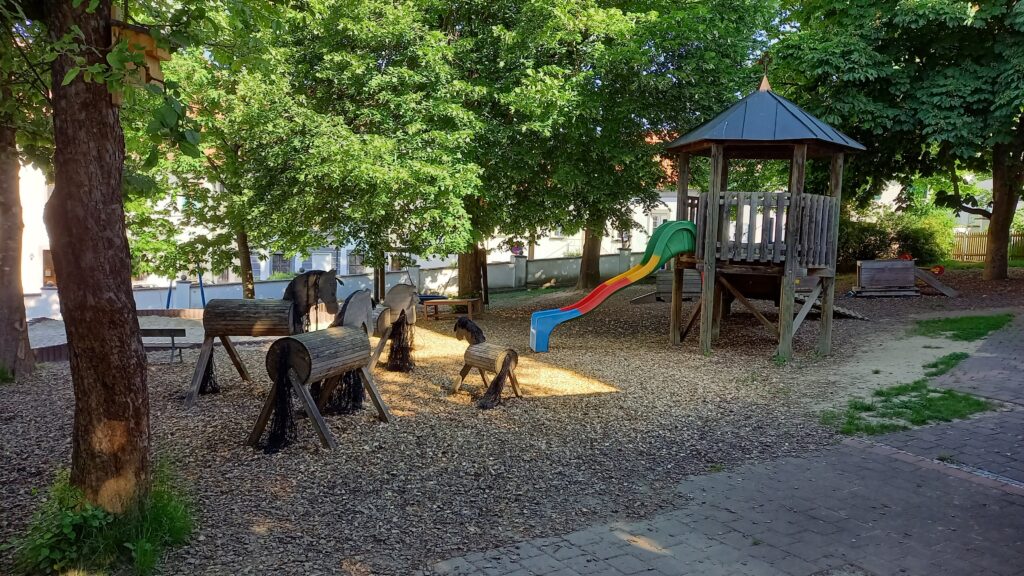 Kinderspielplatz mit Pferden und Rutsche im Park, umgeben von Bäumen.