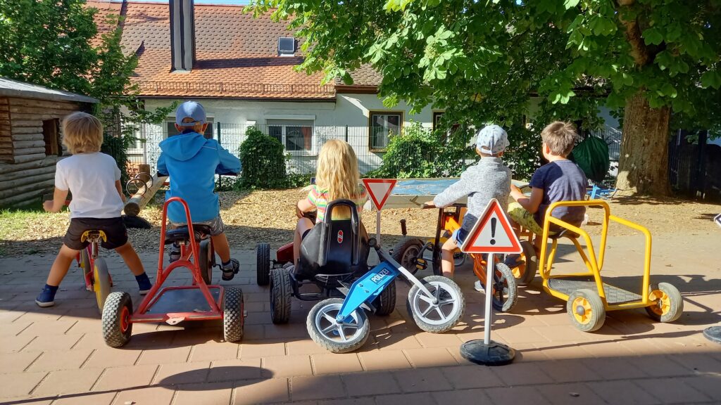 Fünf Kinder mit Spielzeugfahrrädern auf einer Spielplatzstraße im Sonnenlicht.