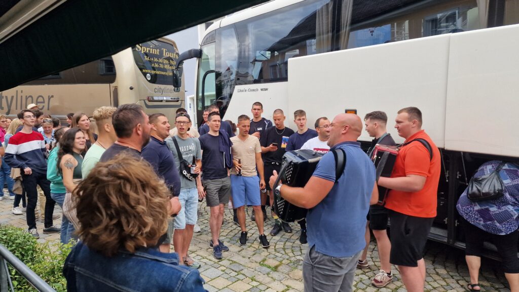 Gruppe von Menschen vor Busse auf einem gepflasterten Platz, einige tragen Akkordeon.