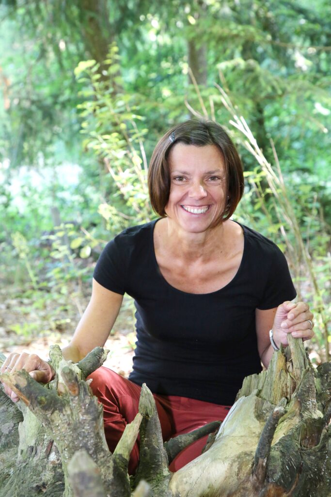 Frau sitzt auf Baumstamm im Wald, lächelt.