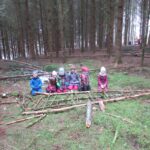Gruppe von Kindern in einem Wald mit Holzstämmen und Stroh auf dem Boden.