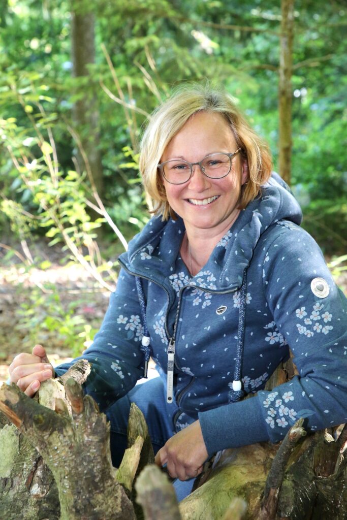 Frau in Wald, lächelnd, mit Holzstück im Schoß.