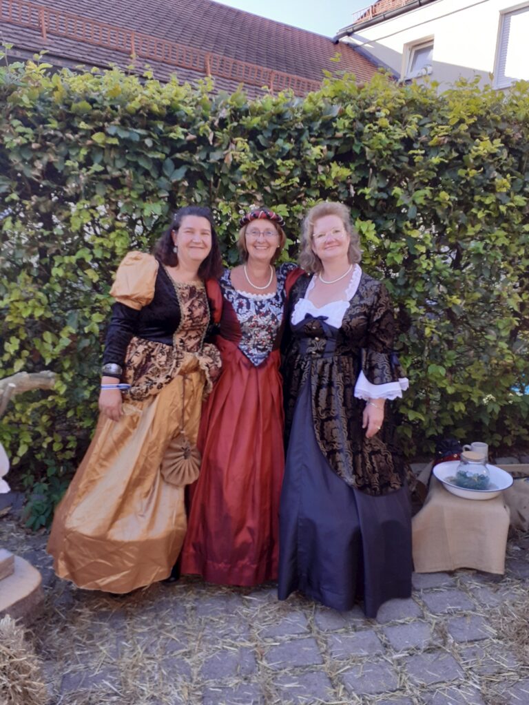 Drei Frauen in historischen Kostümen posieren vor einem grünen Hintergrund.