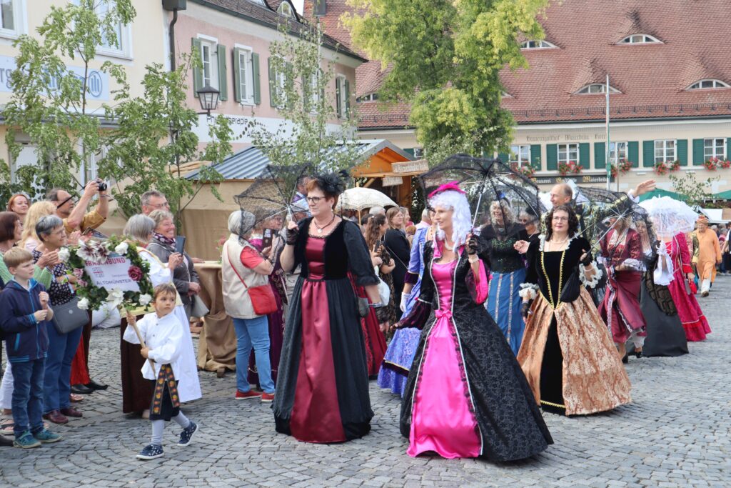 Markt Altomünster: Menschen in historischen Kostümen auf einem gepflasterten Platz, umgeben von Gebäuden und Bäumen.