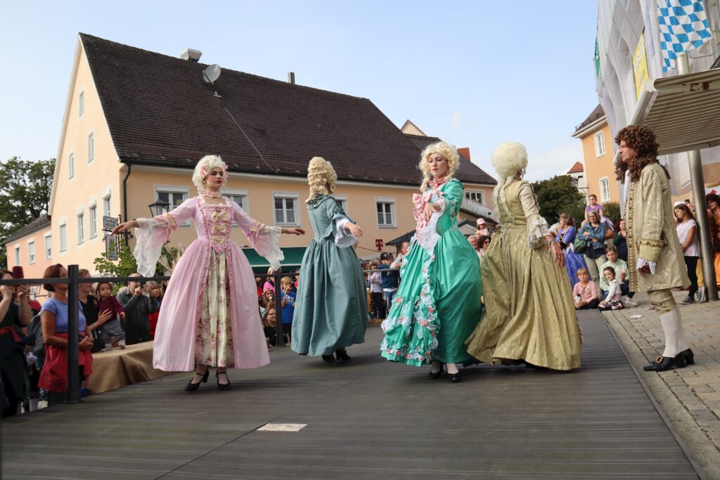 Mädchen in historischen Kostümen auf einer Straße, teilweise in Bewegung, mit Zuschauern im Hintergrund.