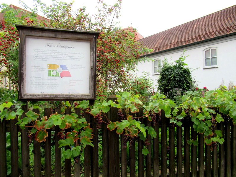 Küsterkräutergarten, Anzeige mit Pflanzen und Zaun vor einem Haus in Altomünster.