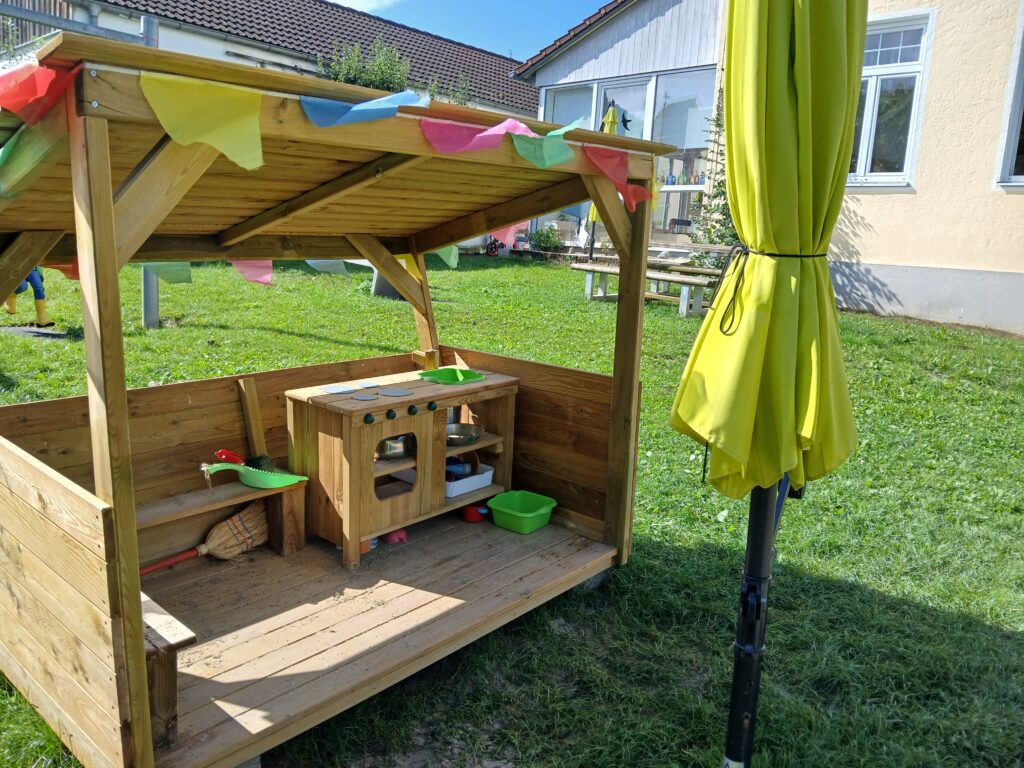 Kinderspielküche im Garten mit Holzstruktur und Sonnenschirm.