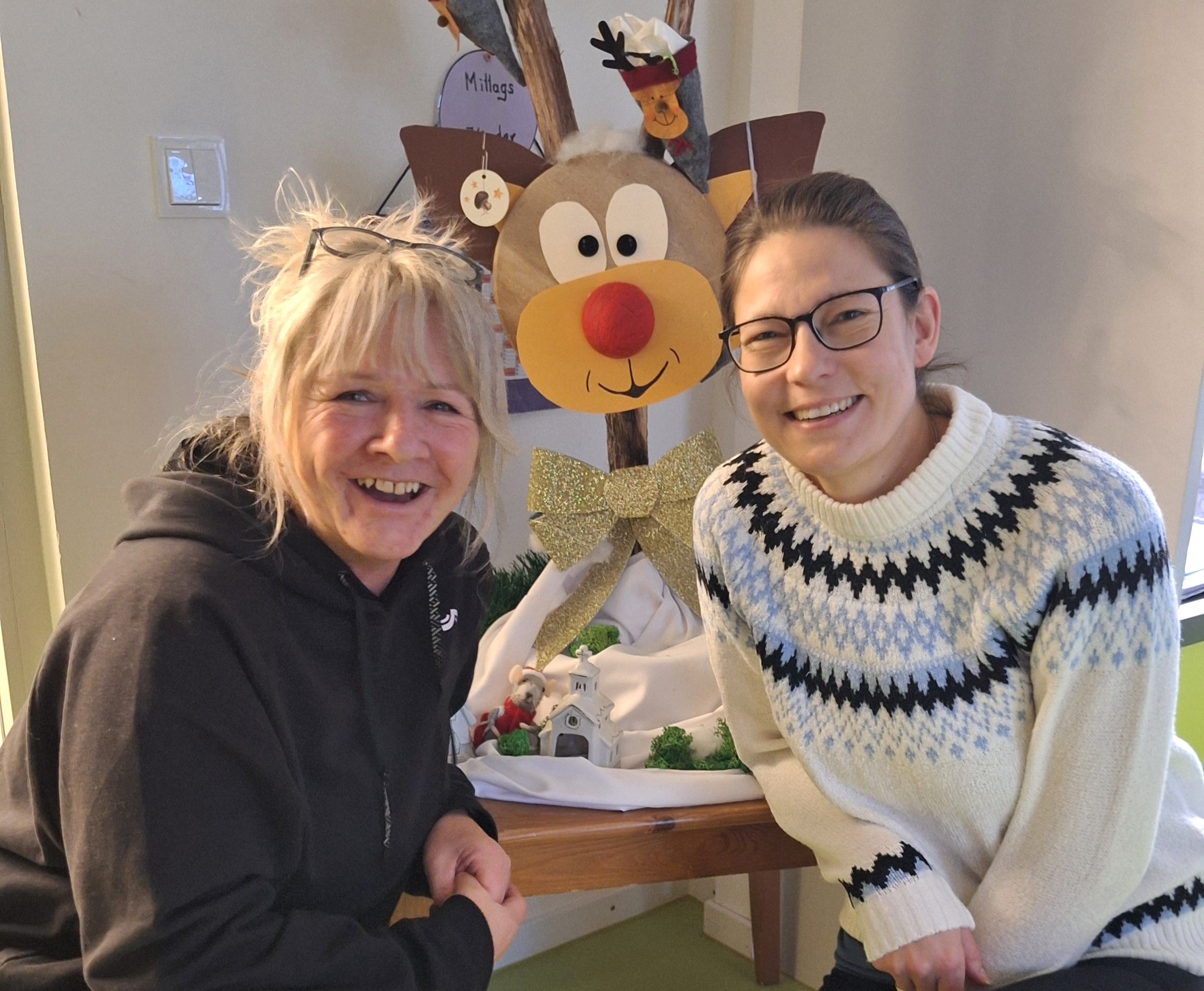 Igel 2025 Zwei Frauen lächeln vor einem Weihnachtsdekoration mit Rentier und Hausmodell.