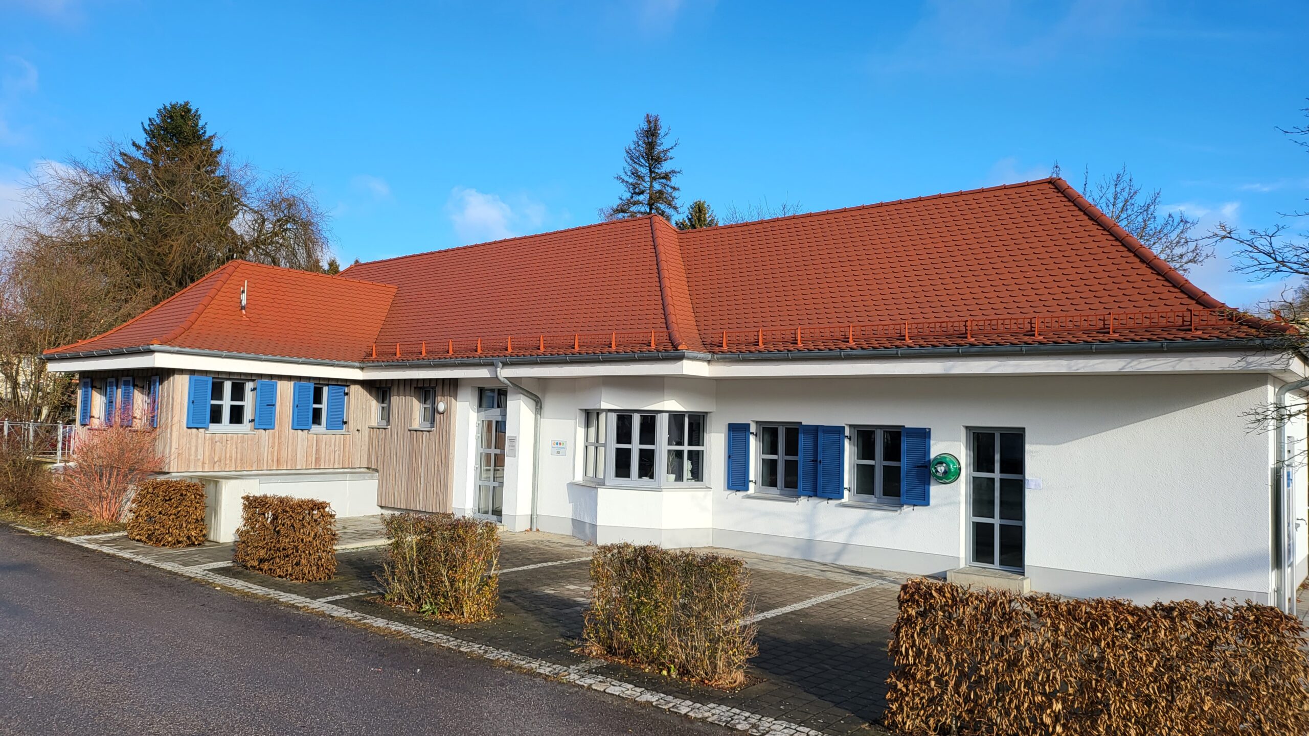 Bauwerk mit rotem Dach und blauen Jalousien. Vor der Fassade sind gepflegte Grünanlagen.