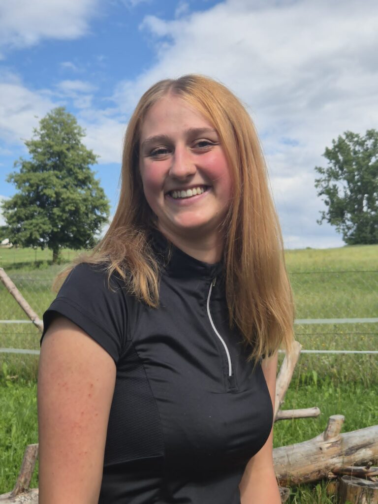 Frau mit blonden Haaren im Freien, lächelt zu den Bildschirmen.