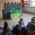 Kinder schauen auf Bühne mit Papierbuden und Puppen in einem Schulraum.