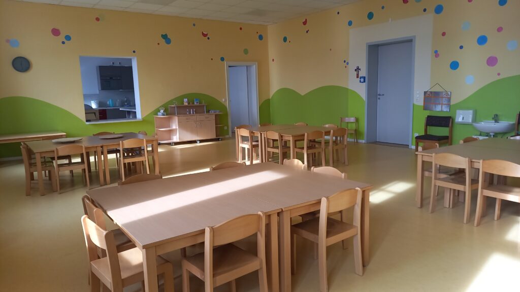 Kindergartenraum mit Tischen, Stühlen und farbenfrohen Wänden.