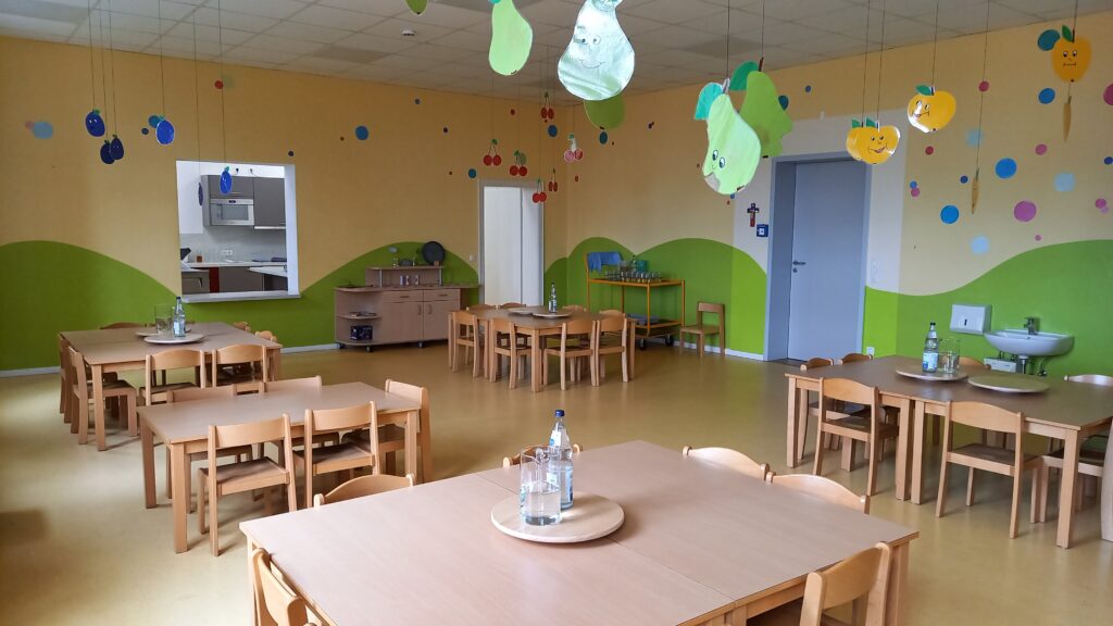 Kindergarten-Räumlichkeiten mit Tischen, Stühlen und leuchtenden, schwebenden Pflaumen- und Äpfel-Ikonen.
