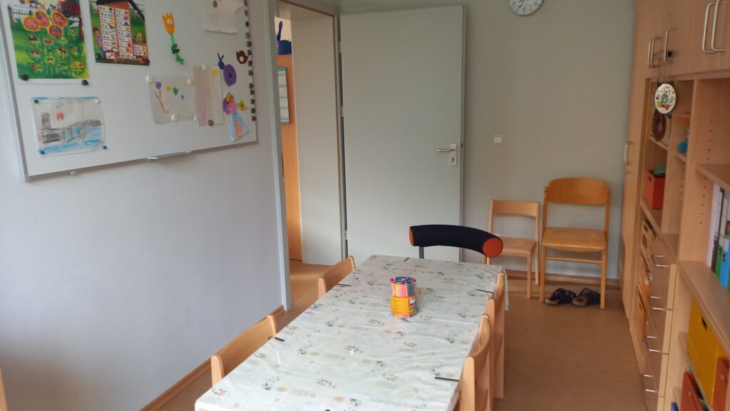 Schülernzimmer mit Tisch, Stühlen und Bildern an der Wand.