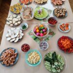 Tisch mit buntem Obst- und Gemüsebuffet, Fisch, Käse, Salat und gekochten Speisen.