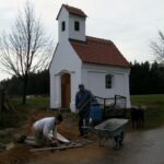 Ein kleines weißes Gotteshaus mit rotem Dach, in dem zwei Männer bei der Ausarbeitung der Umgebung arbeiten.