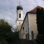 Kirche mit Turm und Schrein auf einem Hügel, umgeben von Bäumen.