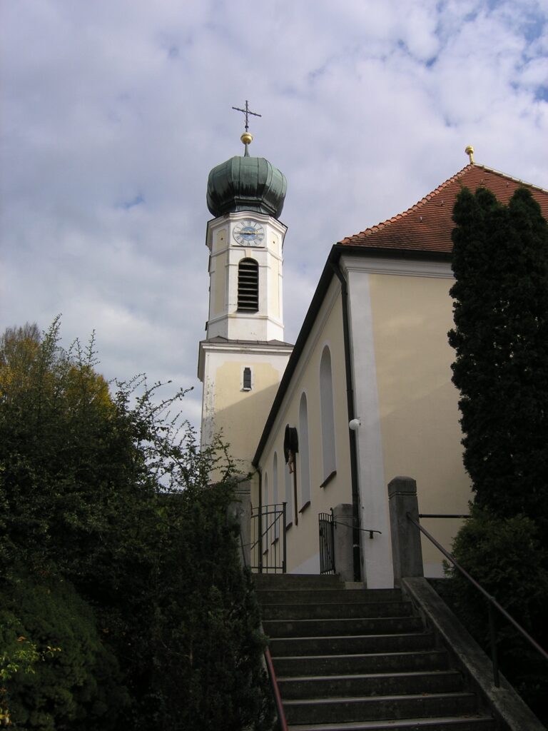 Kirche mit Turm und Schrein auf einem Hügel, umgeben von Bäumen.