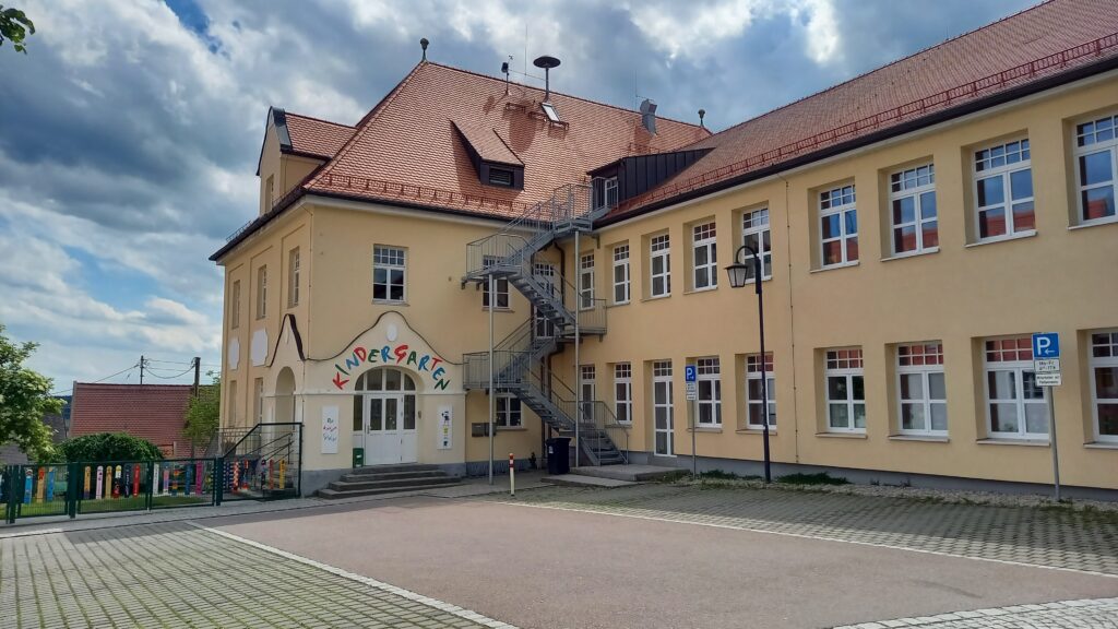 Kindergarten mit gelbem Gebäude, rotes Dach, Stufen und Parkplatz.