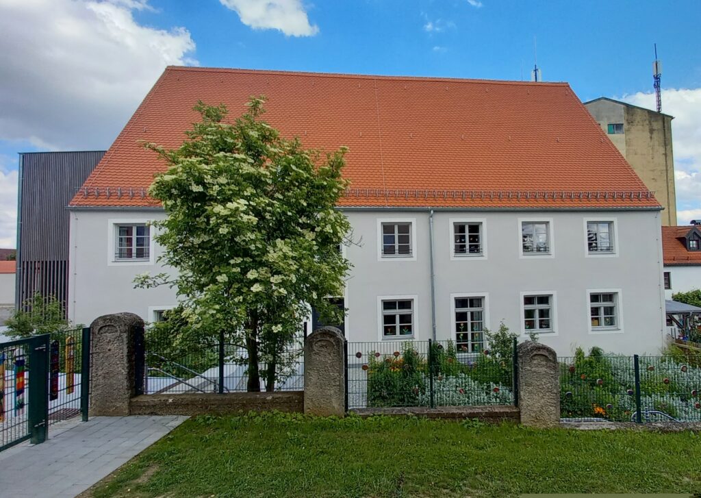 Schule im Marktplatz, rotes Dach, weiße Fassade, gepflasterte Fußgängerzone, grüner Garten mit Kiste und Blumen.