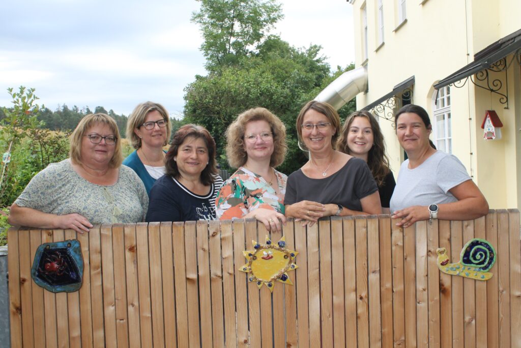 7 Frauen vor einem Zaun mit Kacheln und Garten.