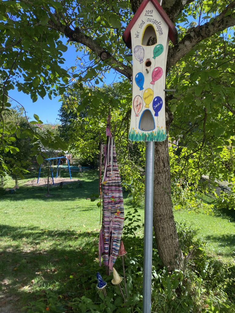 Ein lebendiger Garten mit einer bunten Vogelhütte und dekorativen Objekten im Baum.