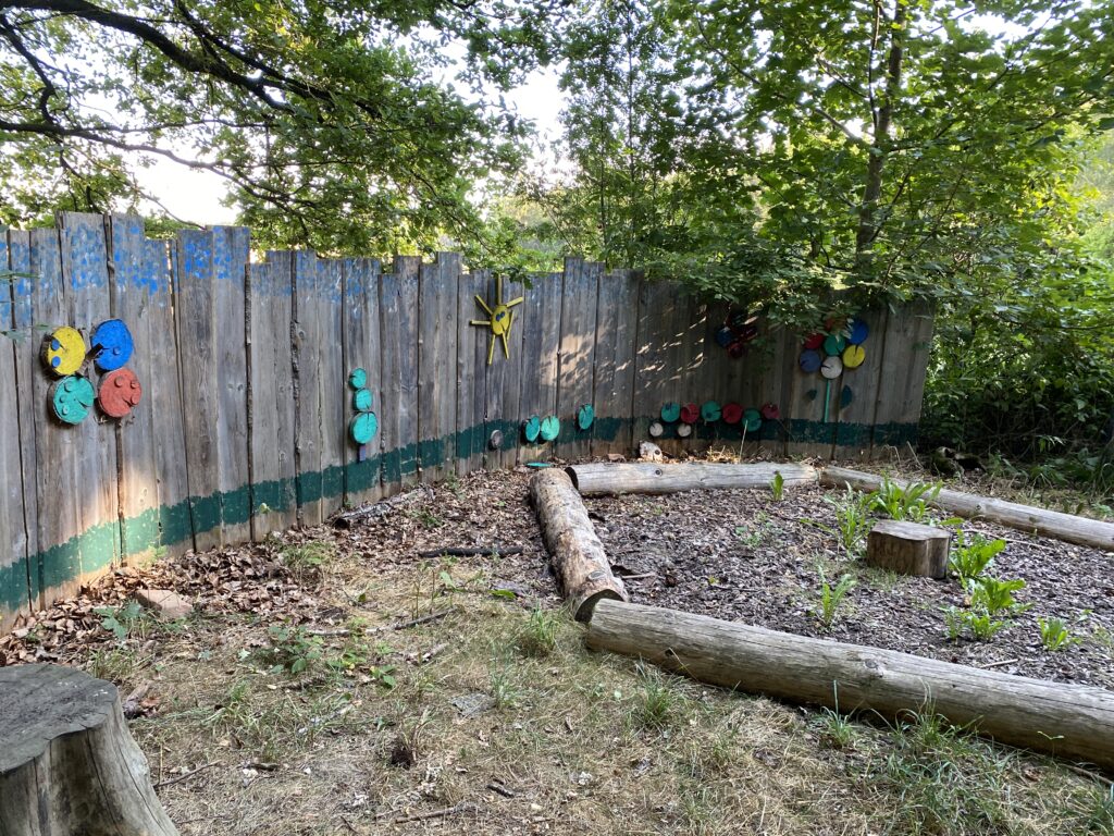 Farbige Spielgeräte an einer Holzumrandung im Park, mit Bäumen im Hintergrund.