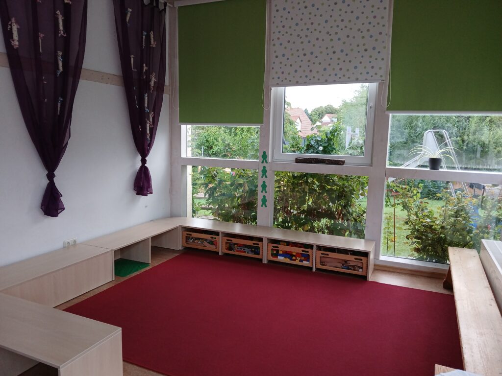 Bild des Kindergartens mit Spielzeugregalen und Fenstern zur Außenansicht.